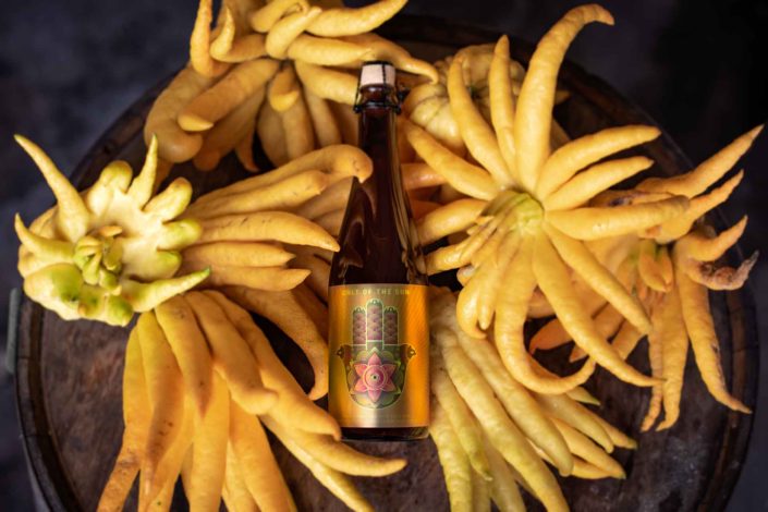 A bottle of Cult of the Sun Sour Blonde Ale resting on a pile of Buddha's Hand Citron