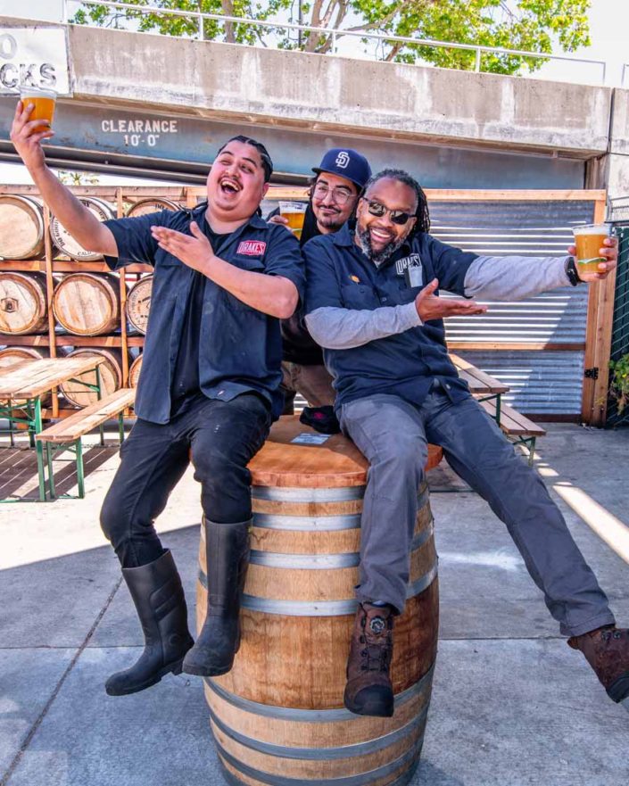 Three men holding a beer sitting on a berr barrel